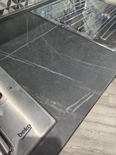 Mira keuken – Moderne rechte keuken met houtlook Zwart aanrecht met rvs spoelbak en kranen, lichte krassen zichtbaar.