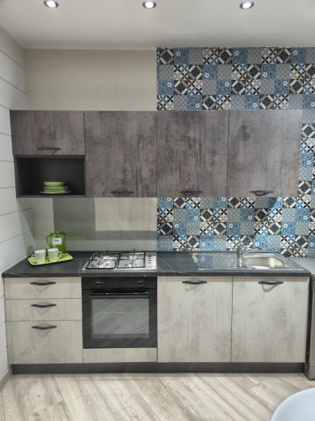 Mira keuken – Moderne rechte keuken met houtlook Moderne keuken met grijze kasten en kleurrijke tegelwand. Gasfornuis en groene accessoires.